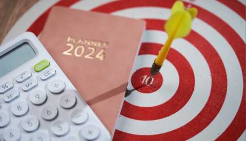 A dart that has hit the target on a dart board is displayed next to a 2024 planning agenda book and a calculator