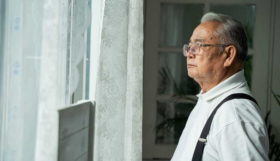Elderly man with glasses wearing suspenders looking out of his window with white curtains