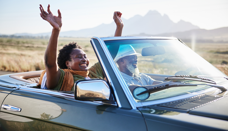 a woman puts her hands up as a man drives her in a convertible on a summer road trip