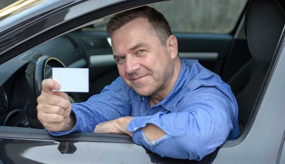 a smiling man in a blue shirt holds his drivers license while leaning out of a window