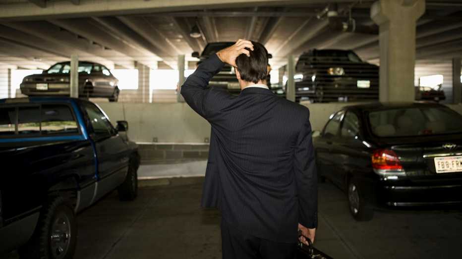 a man in a suit with a briefcase scratches his head while looking for his car in a parking lot