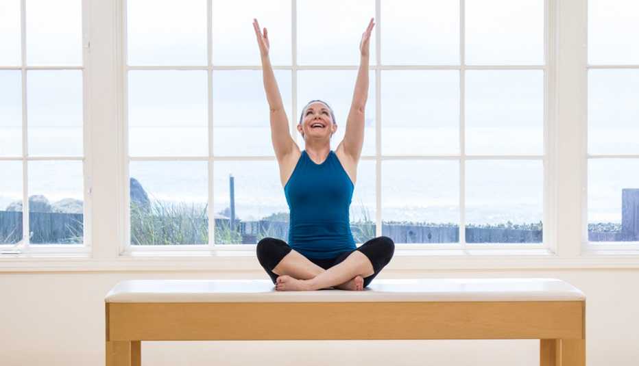 woman doing Pilates, arms raised and legs crossed, on a table in front of a window