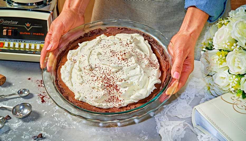 a chocolate cream pie held over a table with measuring spoons, an old blender, white rose bouquet and photo album