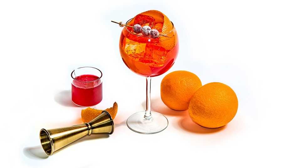 stem glass of cranberry aperol spritz, two oranges, a very small glass of cranberry simple syrup and a brass jigger