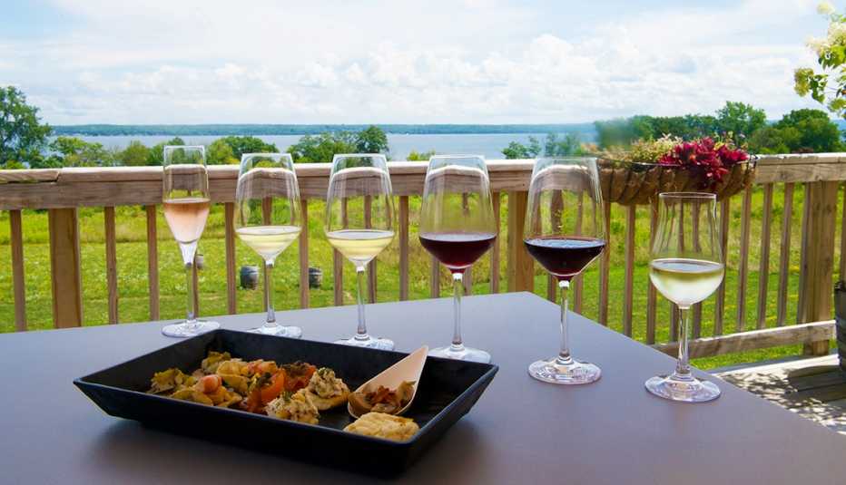 six wine glasses partly filled with red and white wines and a dish of appetizers on a table, overlooking trees and a lake in the distance
