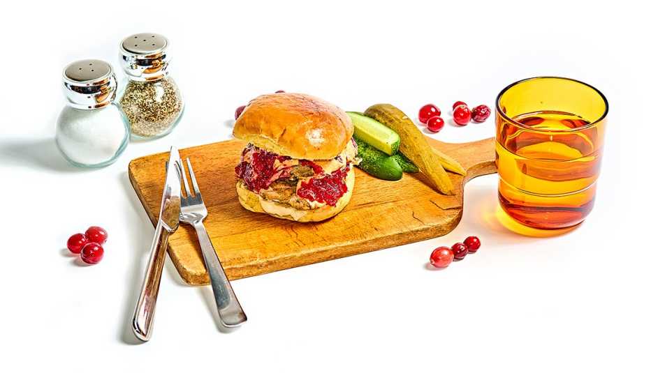 gobbler sandwich and pickles on a wood cutting board, with salt and pepper, knife and fork, glass of water and a few loose cranberries