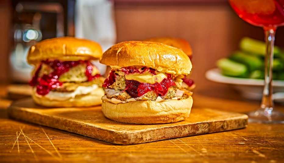 three gobbler sandwiches on buns on a cutting board sitting on a wood table with a stem glass with a pink drink and a plate of pickles