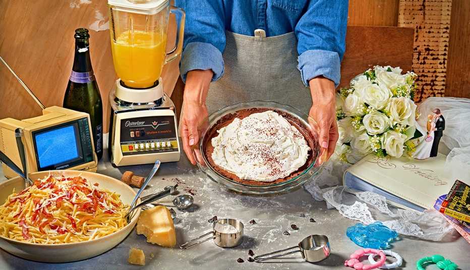 someone wearing an apron holding a chocolate cream pie over a table with a bowl of spaghetti carbonara, parmesan cheese wedge, measuring spoons, an old blender with orange juice, champagne bottle, white rose bouquet, wedding figures, photo album, baby rattles and tiny old TV