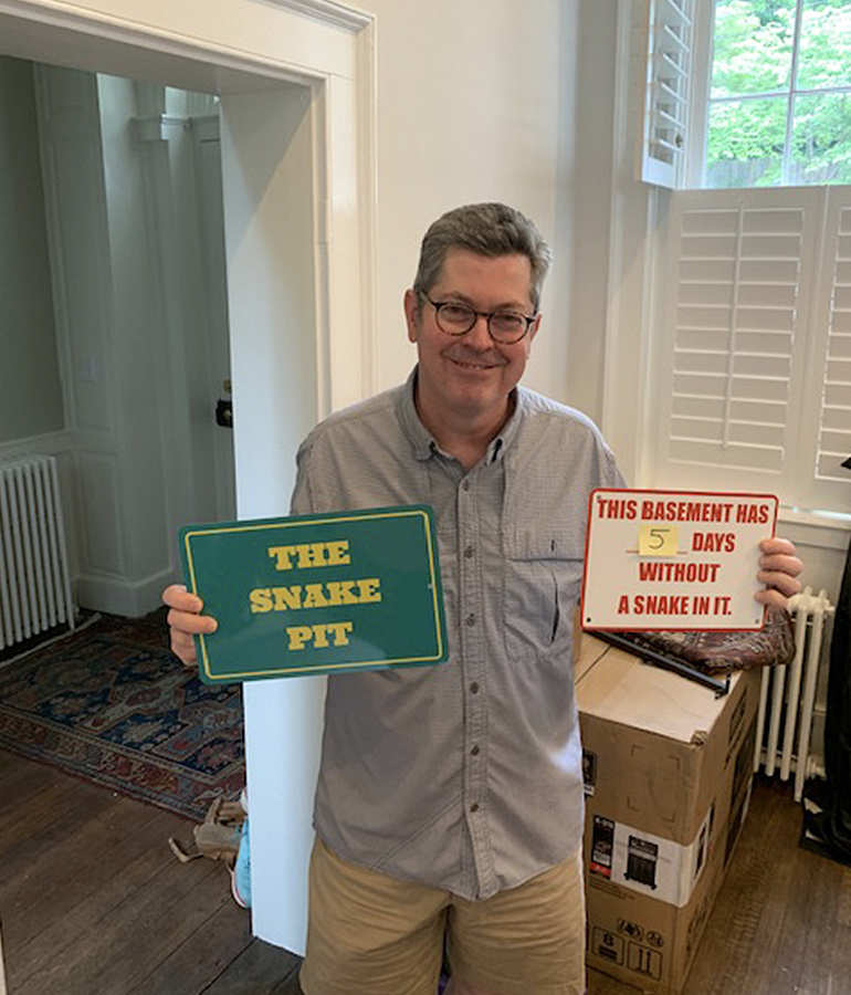 paul schorn holds up two signs inside a house; the green sign says the snake pit and the white one says this basement has 5 days without a snake in it