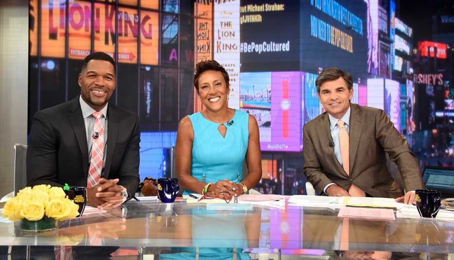 michael strahan, robin roberts, and george stephanopoulos sit at desk in front of window that shows part of downtown new york city