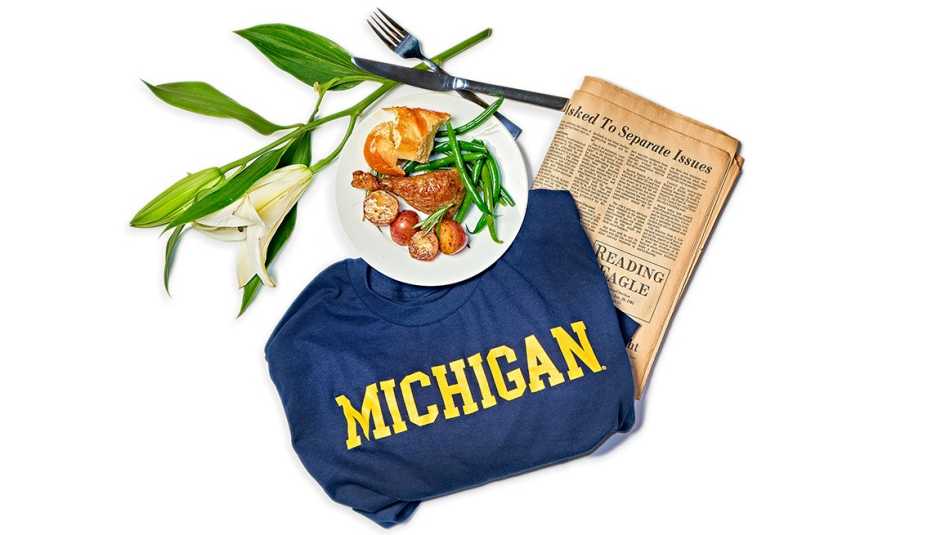 Michigan t-shirt, yellowed newspaper, white flower, and plate of chicken with green beans, potatoes, and bread and fork and knife