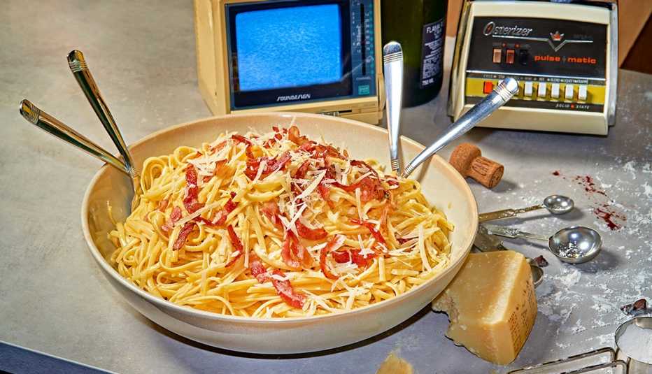 bowl of spaghetti carbonara with four utensils sticking out, on a table with a wedge of parmesan cheese, an old blender, measuring spoons, a cork and a tiny old TV