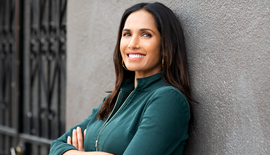 padma lakshmi smiling with arms crossed and leaning against a gray wall 