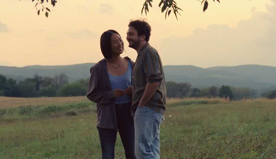 greta lee as nora and john magaro as arthur standing in grassy field with mountain in the distance in still from past lives