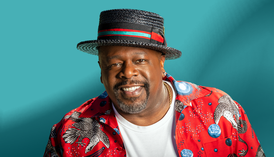 cedric the entertainer in red shirt and dark hat with red and blue ribbon; blue background