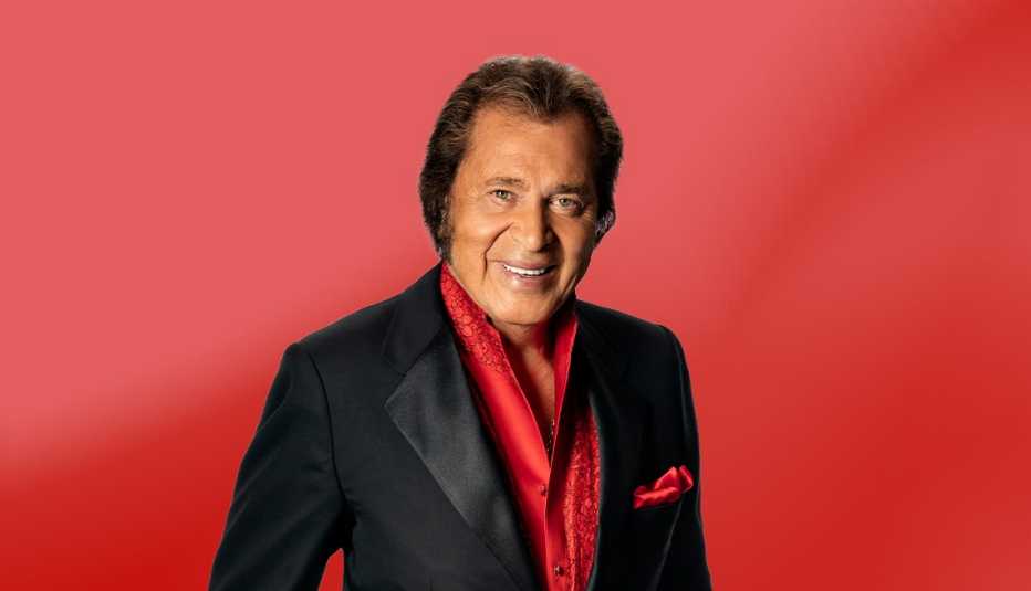 engelbert humperdinck wearing black suit jacket with red button down shirt underneath, against red ombre background