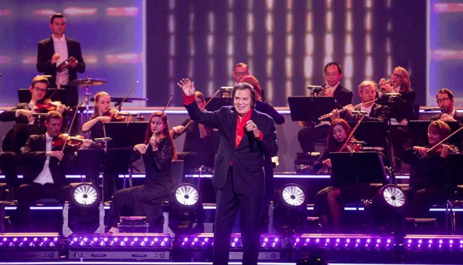 Engelbert Humperdinck performing on stage, waving to crowd, with people behind him playing different instruments