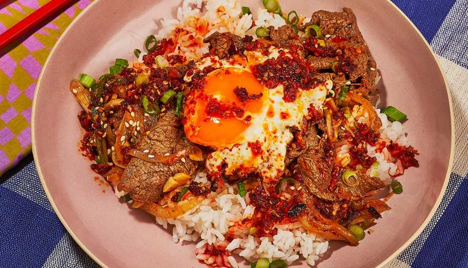 bowl of chili crisp bulgogi deopbap from chili crisp cookbook, showing fried egg with exposed yolk atop a bowl of rice and sliced beef with chili sauce and green opions