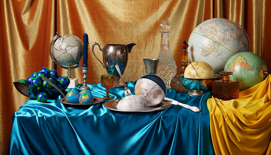stylized table setting featuring shiny blue and gold cloths and world globes of varying sizes and designs, some on plates with utensils and some in bowls, along with a candle, brass water pitcher and ornate glass wine decanters