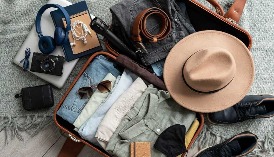 open suitcase with clothes, hat, belt and sunglasses in it; shoes, laptop, headphones and camera are lying next to it