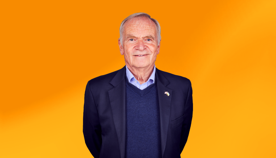 jeffrey archer wearing navy blue suit jacket with navy blue shirt against orange-yellow ombre background