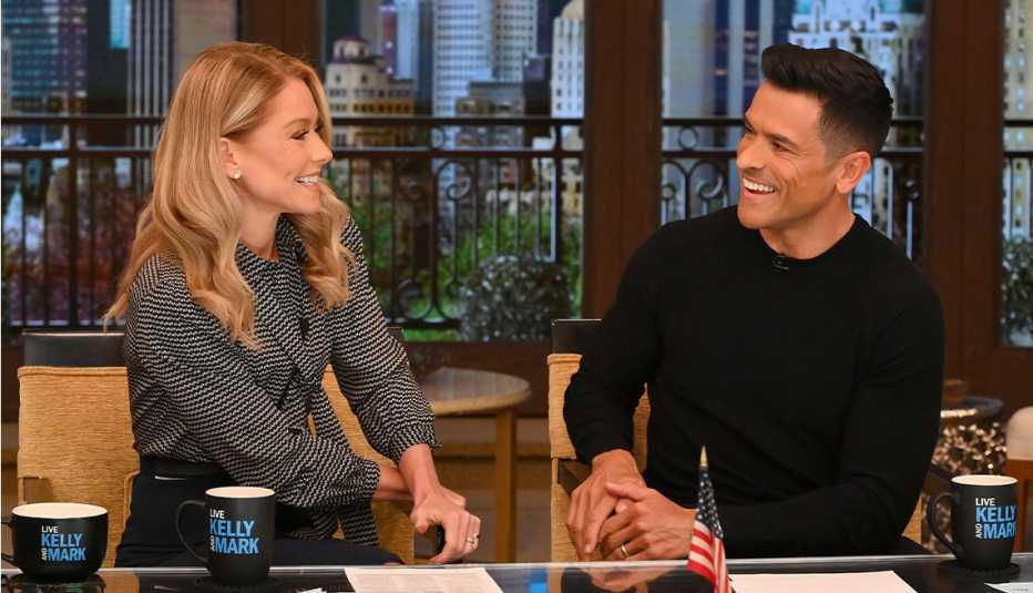 kelly ripa and mark consuelos sitting at desk turned towards each other; mugs that say live kelly and mark are in front of them