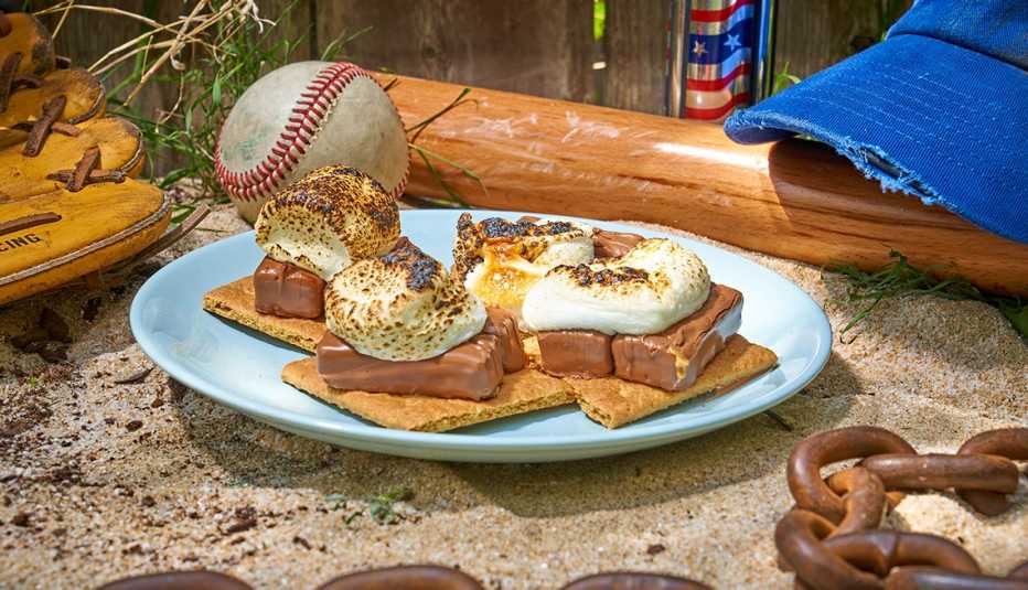 four milky way melt smores on a blue plate on sand alongside a baseball, glove, hat, trophu and rusty chain