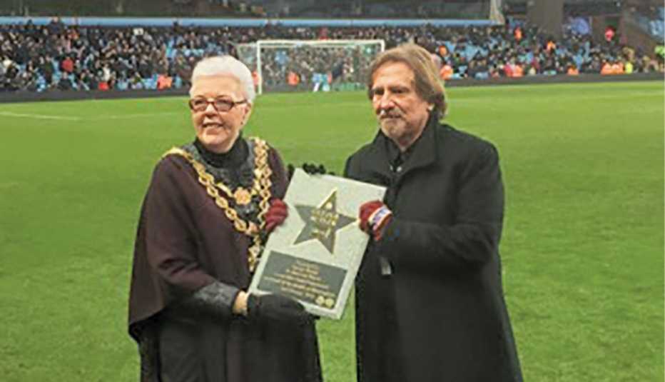 geezer butler receiving the walk of stars award from woman on the field at villa park; crowd in stands in the distance