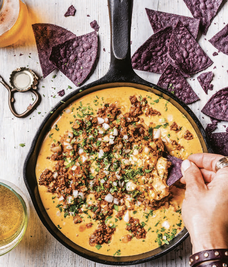 skillet with queso in it, topped with chorizo, onion, chiles; hand dipping tortilla chip in queso; tortilla chips on table around skillet
