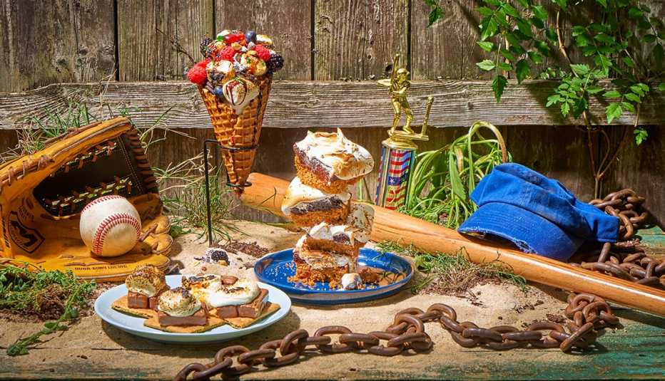 milky way melt smores on a plate, smores bars piled high on a plate, smore parfait overflowing with berries, candy and marshmallows in a metal stand, alongside a baseball in a glove, bat, hat trophy and rusty chain, all on sand in front of a wood fence