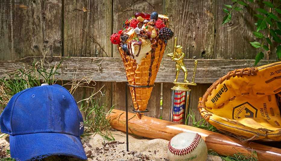 smore parfait in a waffle cone held by a metal stand overflowing with berries, toasted marshmallows and candy, alongside a baseball,  glove, blue baseball hat and bat, all on top of sand