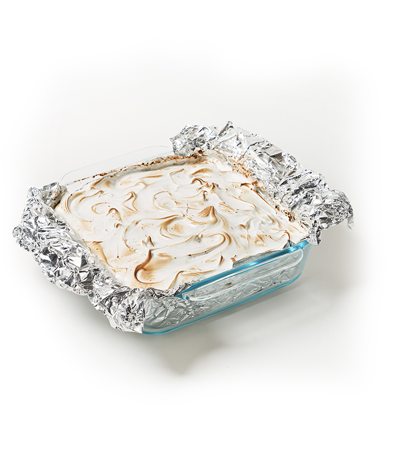 smores bars topped with meringue in glass dish with aluminum foil over two sides