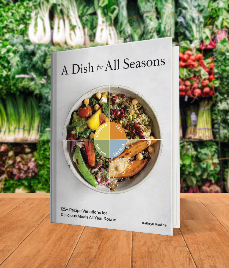 cookbook that says a dish for all seasons with bowl showing a different salad in each quadrant for summer, fall, winter and spring; on wooden table with vegetables and plants behind it