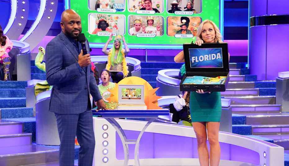 wayne brady holding microphone; tiffany coyne holding open case that says florida; a few people in background in person and on a screen on the set of let's make a deal 