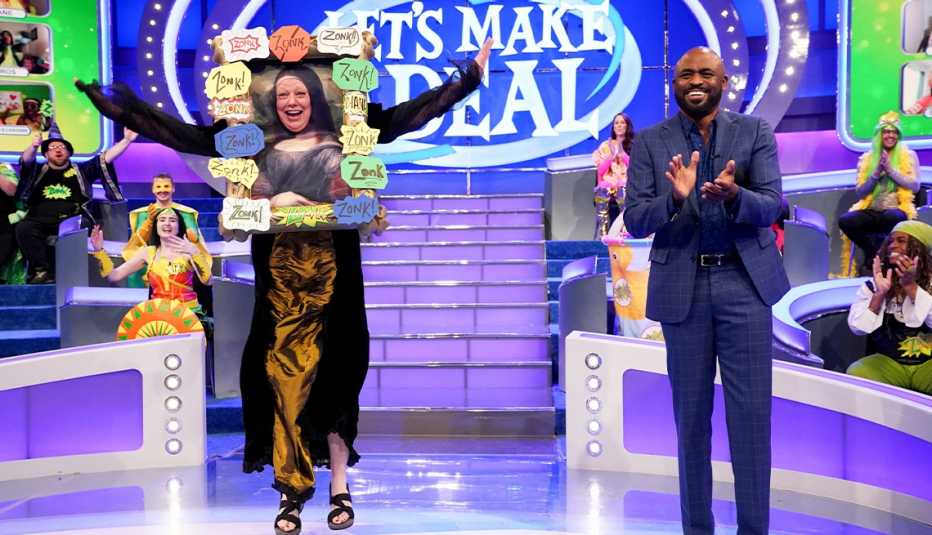 wayne brady clapping next to person wearing a frame that says zonk all around it on set of let's make a deal; contestants behind brady; giant screen in background that says let's make a deal