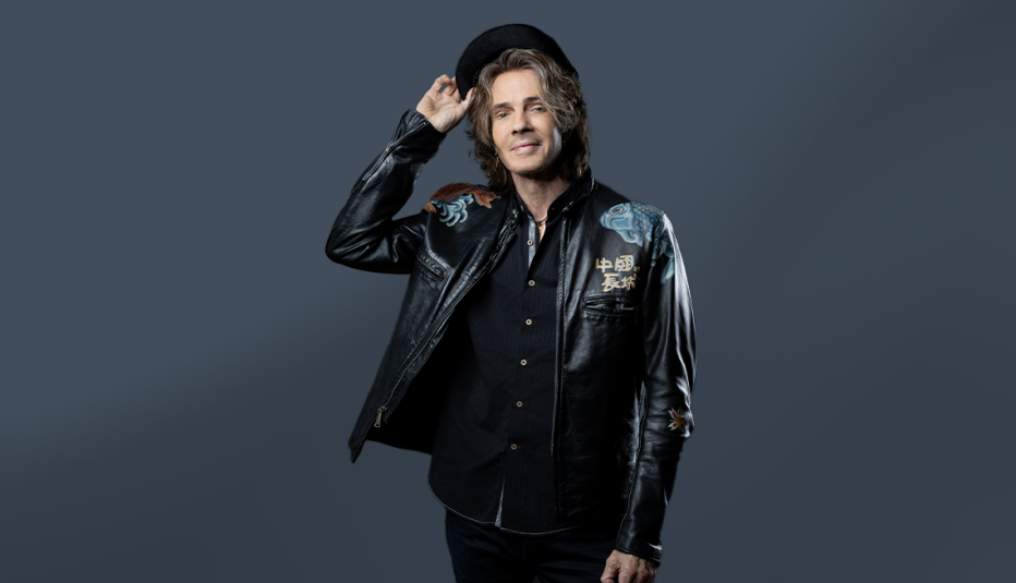 rick springfield in all black, holding hat tipping off head; gray background