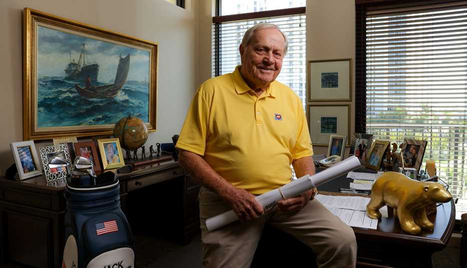 Jack Nicklaus leaning on desk, holding 2 rolled up pieces of long paper; golf club set next to him; pictures and globe in the background