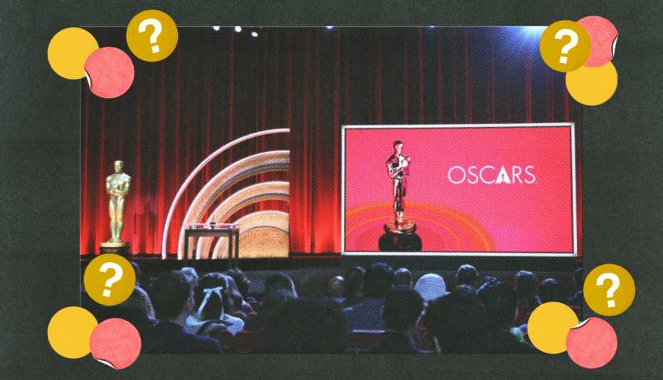 wide shot of oscars stage with statuette and word oscars on screen on right and another statuette on left; people in the audience; yellow, pink and gold circles with question marks in four corners