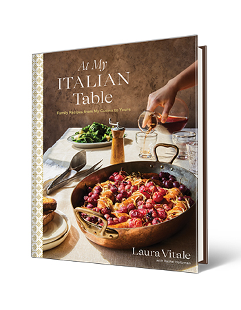 Book cover that says At My Italian Table Family Recipes from My Cucina to Yours, Laura Vitale; pan and plates with food on it on a table