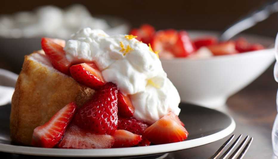 piece of strawberry shortcake on plate; bowl of strawberries behind it
