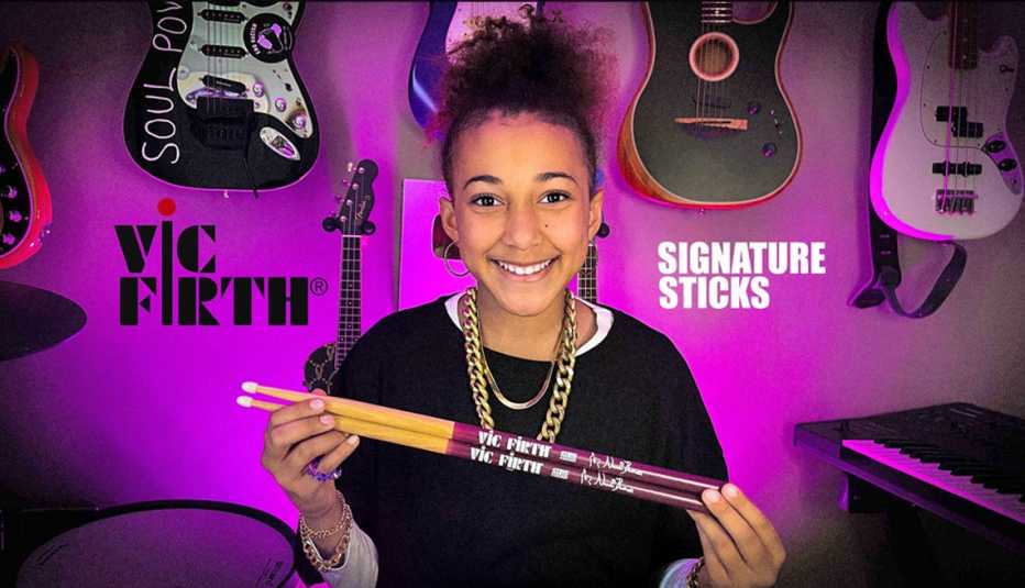 Nandi Bushell holding two drum sticks with guitars, a keyboard and the words Vic Firth and Signature Sticks behind her
