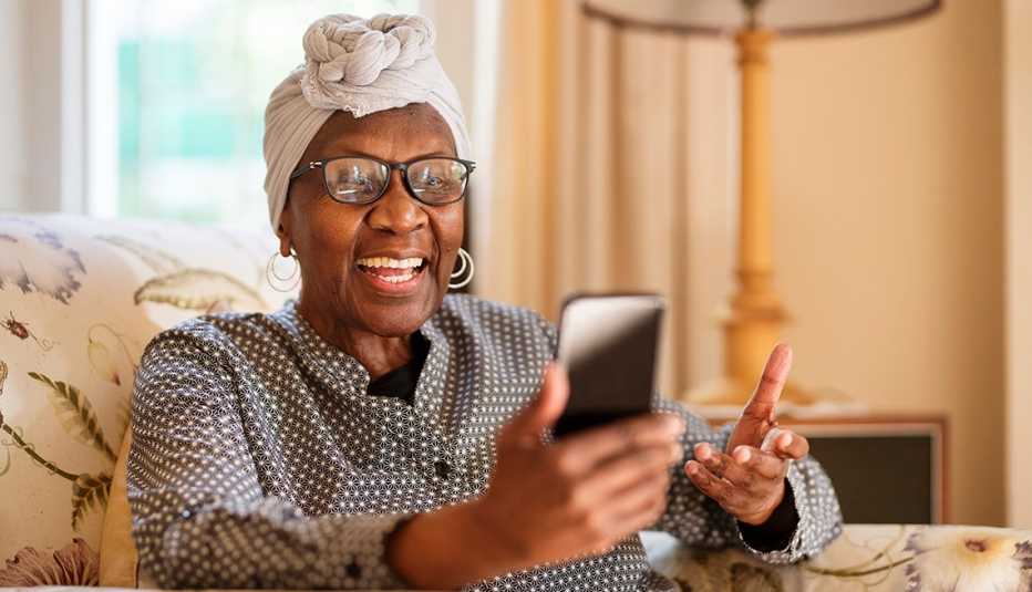 A senior woman smiles in a chair at home while video calling on her smartphone.