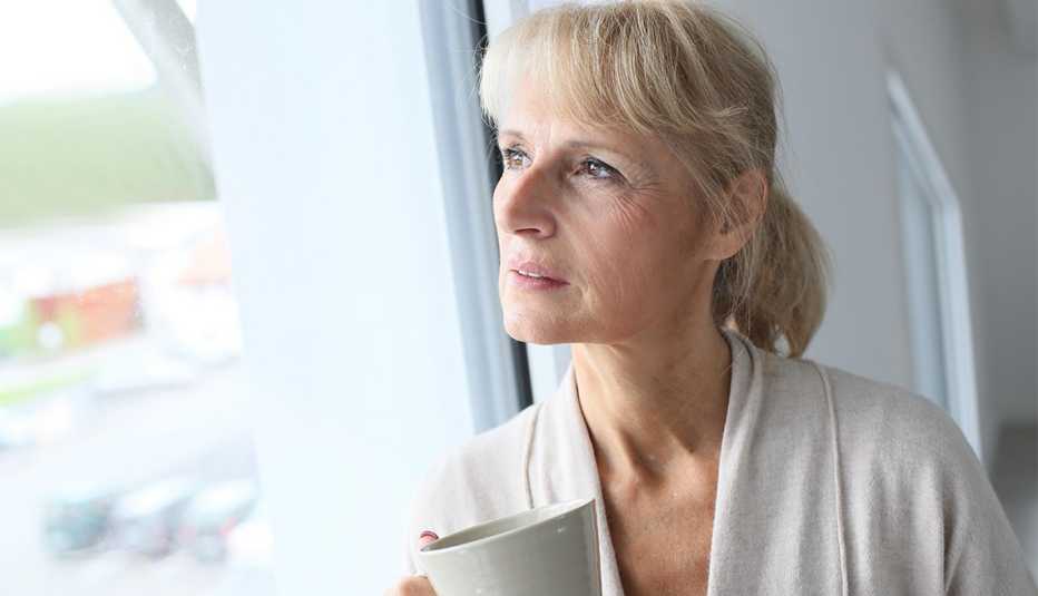 a lonely looking woman holding a coffee mug looking out a window