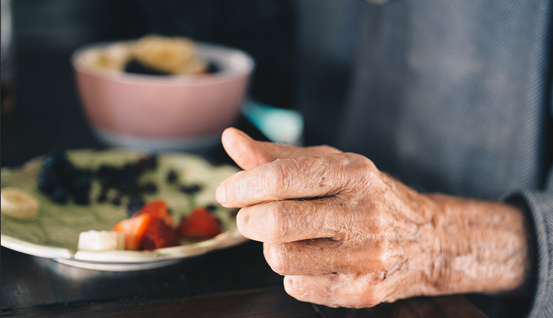 Hand of an elderly man who is sitting at a table with a bowl of fruit and cereal