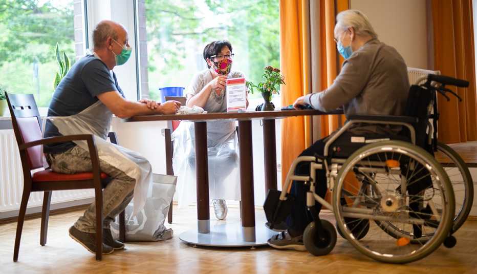 a couple and a man in a wheelchair sitting around a table in a nursing home wearing personal protective gear