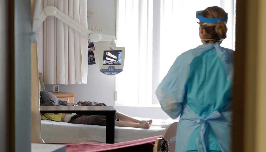 A medical worker wearing personal protective gear stands in the doorway of a nursing home room as a patient lays in bed inside