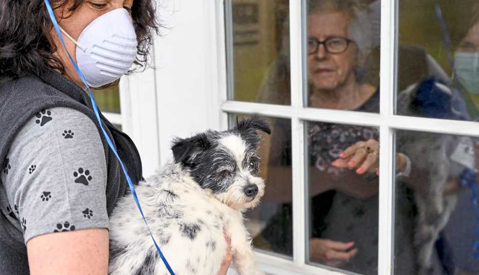 Woman wearing a mask holding a small dog visiting a woman through a nursing home window
