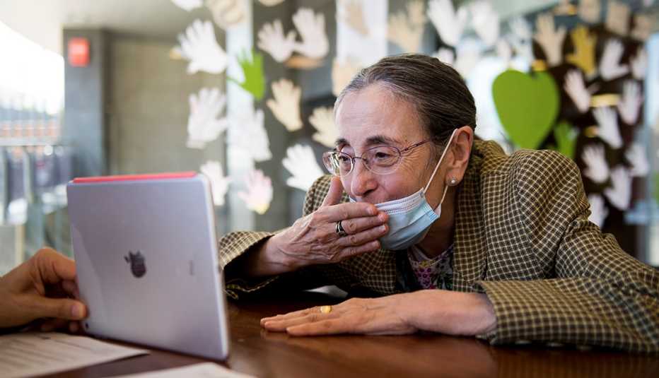 woman in a nursing home wearing a mask and blowing a kiss into a tablet that is being held by a care worker