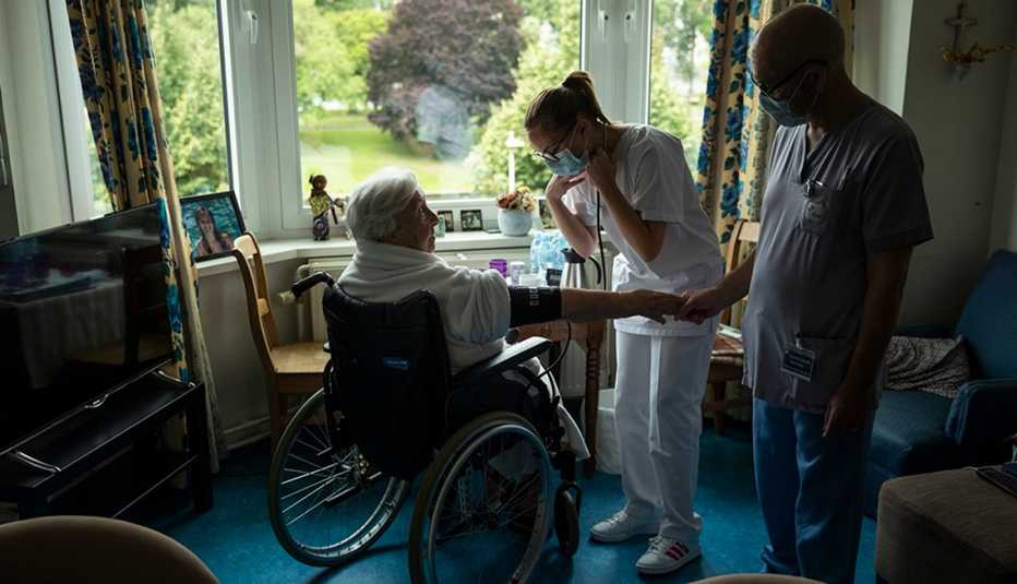 A woman in a wheelchair sits in her nursing home room being attended two by two nursing home staff members wearing face masks