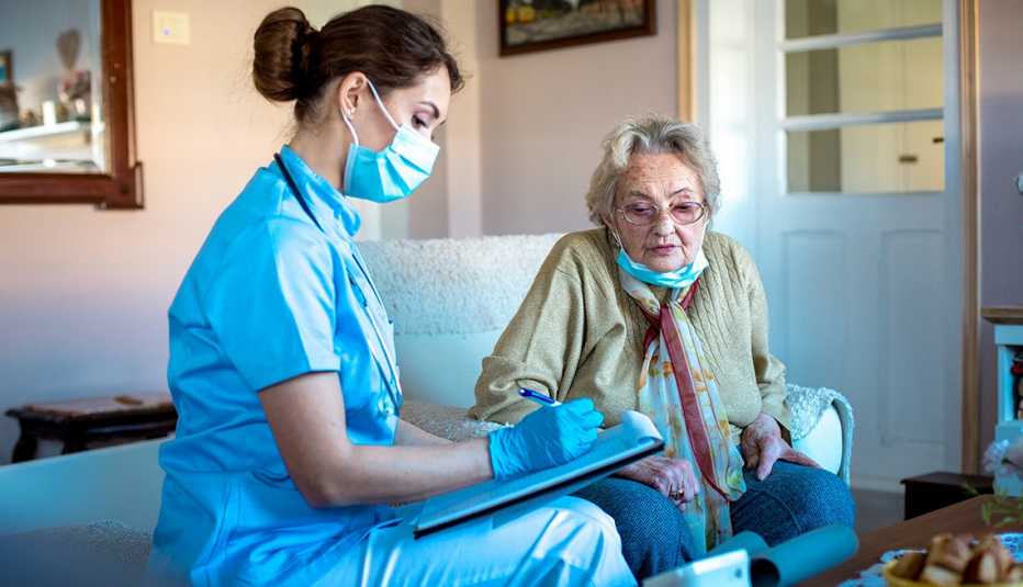 Adult woman is visited by district nurse from home care. Nurse is holding her clip board, writing down patient's information. They are in living room, sitting and talking.